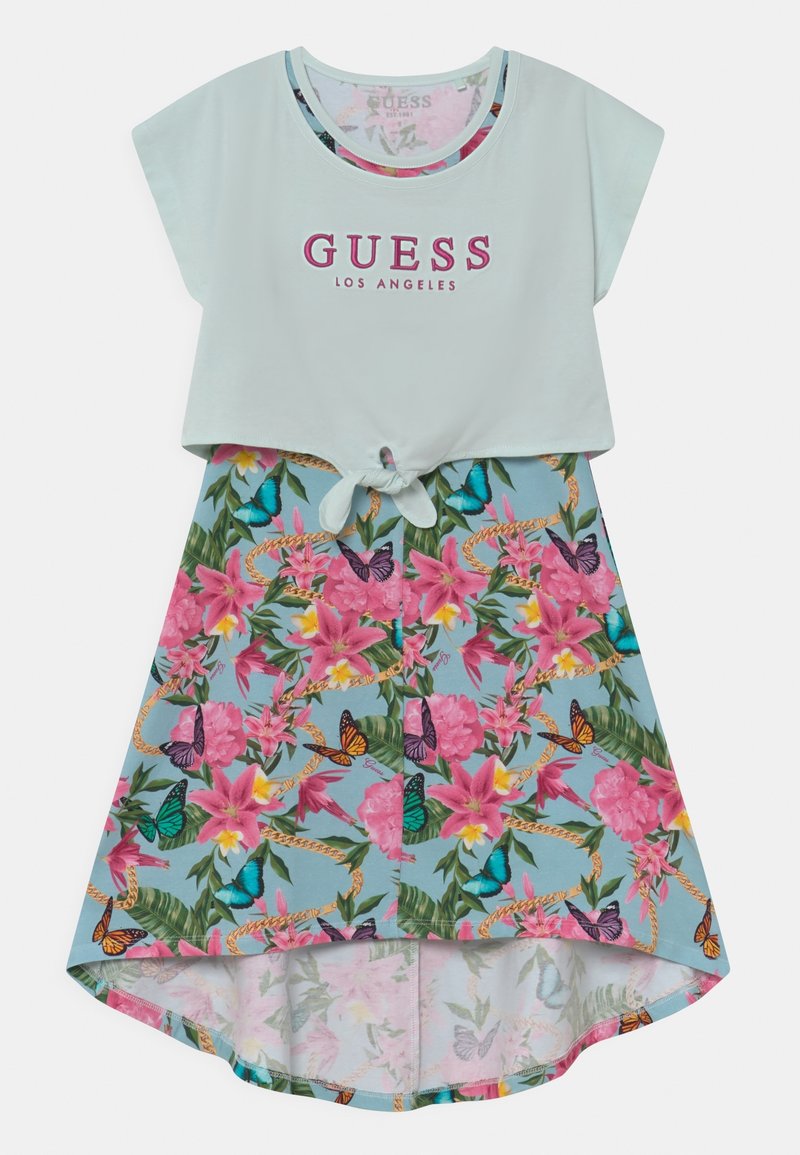 Top court blanc avec le texte « GUESS Los Angeles » noué à la taille sur une jupe courte devant et longue derrière avec un fond bleu, des fleurs roses, des papillons et des feuilles vertes.