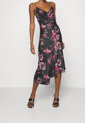 Mujer que lleva un vestido midi de satén negro con estampados rosas de flores y frutas y tirantes finos, combinado con sandalias negras de puntera abierta.