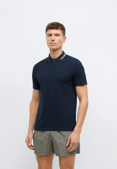 Tamsiai mėlynas polo marškinėlis iš lygaus audinio su trijų mygtukų apykakle ir kontrastingu apykaklės akcentu; derinamas su dryžuotais šortais.