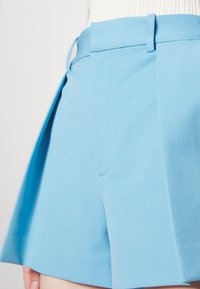 Shorts bleu clair sur mesure avec passants de ceinture, portés avec un haut blanc côtelé, montrés de la taille au milieu de la cuisse sur un fond clair.