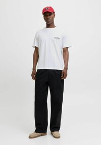 White cotton t-shirt with short sleeves and small graphic. Black wide-leg trousers. Beige slip-on shoes. Red cap with logo detail.