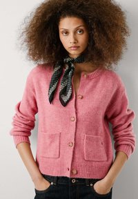 Een jonge vrouw met krullend haar draagt een roze cardigan met zakken en een zwart bedrukte sjaal, poserend tegen een neutrale achtergrond.