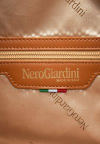 Braune Leder-Reißverschlusszieher vor einem goldenen Satin-Hintergrund. "NeroGiardini" in Gold geprägt über einem Detail der italienischen Flagge. Elegante Nahtstickerei.