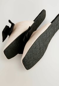 Sandali piatti con suola in gomma nera texturizzata, soletta in beige morbido e dettagli in tessuto nero sulla parte superiore, con un fiocco decorativo.