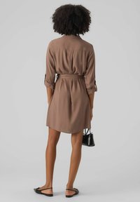 Abito camicia marrone con vita cintata, maniche lunghe arrotolate e vestibilità comoda. Abbinato a sandali neri e una piccola borsa nera.