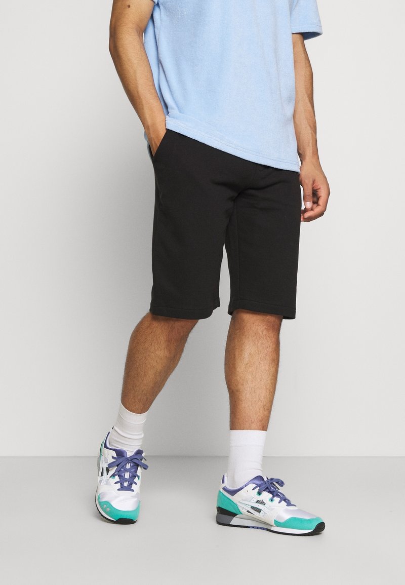 Homme portant un short noir jusqu’au genou, une chemise bleu clair à manches courtes, des chaussettes blanches et des baskets blanches avec des accents turquoise et violets.
