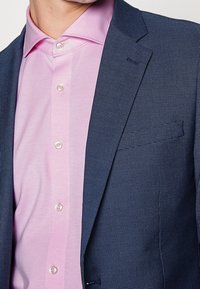 Mørkeblå struktureret blazer med klapkrave, parret med en lys pink skjorte med knapper og en glat overflade.