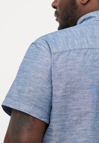Camisa de manga corta en tela azul claro, con un acabado texturizado y un diseño limpio y sencillo. Costuras visibles y detalles en el collar.