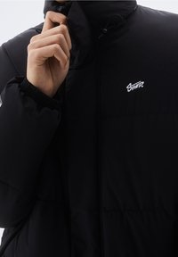 Doudoune noire avec un col montant et des poignets élastiques. Elle présente un petit logo blanc sur la poitrine et une texture lisse et brillante.