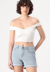 Biela crop top bez ramienok s nariaseným detailom vpredu, skombinovaný so svetlomodrými džínsovými šortkami s ohrnutými manžetami a predným zapínaním na gombík.