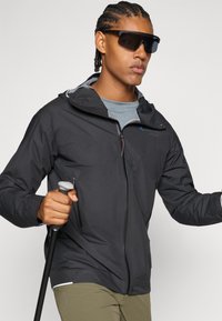 Leichte schwarze Jacke mit Reißverschluss, Kapuze und Netzeinsätzen. Kombiniert mit einem grauen Shirt und einem Trekkingstock, getragen mit olivfarbenen Hosen.