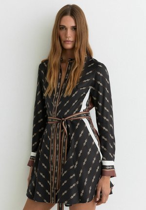 Femme aux longs cheveux bruns portant une robe noire à motifs avec des rayures marron et blanches, debout devant un mur blanc.