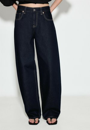 Person iført løstsiddende mørkeblå denimjeans med synlige syninger, kort sort top og sorte sandaler med firkantet tå, der viser tæerne.