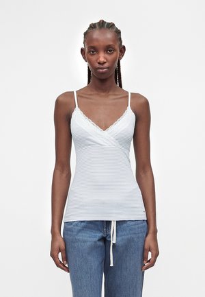 Jonge vrouw met gevlochten haar draagt een witte gestreepte top met kanten rand en blauwe spijkerbroek tegen een effen witte achtergrond.