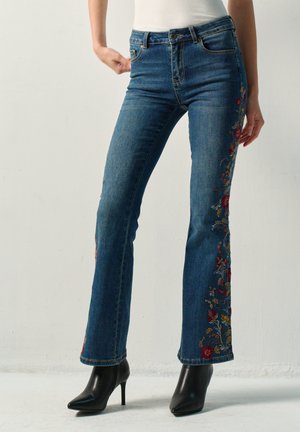 Persona que lleva vaqueros azules de talle alto con bordados florales a los lados, combinados con botines negros de tacón alto y una camiseta blanca.