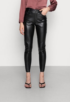 Vrouw die strakke zwarte leren broek, mauve blouse met knopen en zwarte open-toe hoge hakken draagt, staand tegen een effen muur.