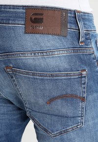 Jeans de mezclilla azul con una etiqueta de cuero marrón que presenta "G-STAR RAW." Costuras visibles en los bolsillos y un acabado descolorido y texturizado.