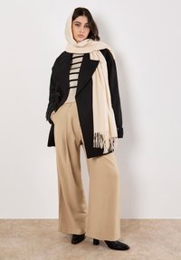 Cappotto nero doppiopetto sopra un maglione a righe crema, abbinato a pantaloni beige a gamba larga e a una sciarpa crema con frange. Stivaletti neri con tacco.