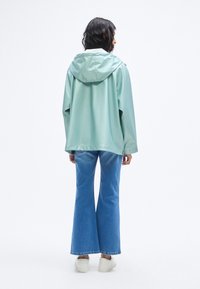 Αδιάβροχο μπουφάν σε μέντα πράσινο με κουκούλα, άνετη γραμμή και κουμπιά πατιλέτα· συνδυασμένο με ανοιχτό μπλε καμπάνα τζιν και λευκά αθλητικά παπούτσια.
