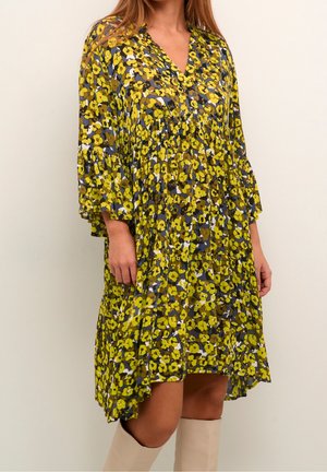 Kvinde iført en løs kjole til knæet med gul, brun og blå blomsterprint og 3/4 længde pufærmer, kombineret med beige støvler.