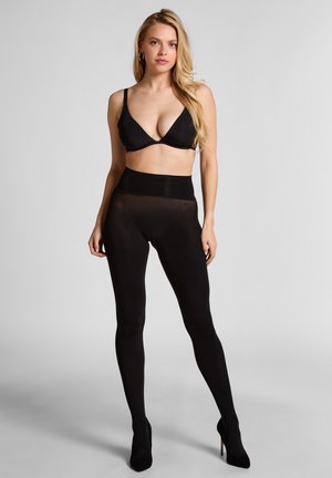 Schwarze dehnbare Leggings mit hohem Bund, transparenter Textur, kombiniert mit einem schwarzen dreieckigen Bralette und getragen mit spitzen Absätzen.