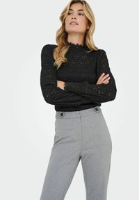 Femme aux longs cheveux blonds portant une blouse noire en dentelle à manches longues et un pantalon gris taille haute, bras croisés, regardant vers le haut.