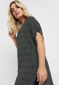 Top nero con pois bianchi, maniche corte, bordi scallop e vestibilità ampia. Presenta un collo rotondo e un orlo posteriore più lungo.
