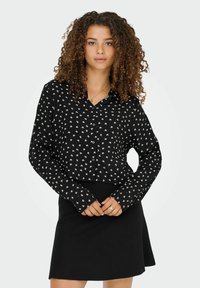 Femme aux cheveux bouclés portant un haut noir à manches longues avec des croix blanches et une jupe noire, se tenant les mains croisées.