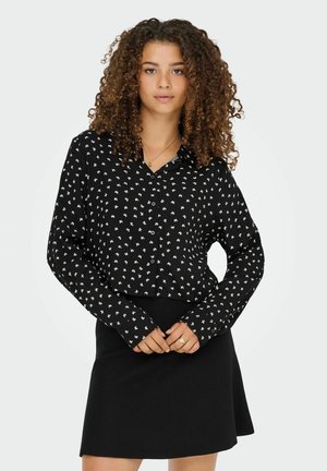 Femme aux cheveux bouclés portant un haut noir à manches longues avec des croix blanches et une jupe noire, se tenant les mains croisées.