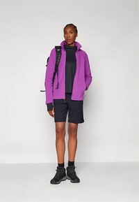 Γυναίκα ντυμένη με μωβ μπουφάν, μαύρες βερμούδες, μαύρες μπότες και μαύρο σακίδιο, στέκεται μπροστά σε απλό γκρι φόντο.