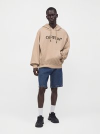 Homme portant un sweat à capuche beige Off-White, un short en jean bleu, des chaussettes blanches et des baskets noires, debout devant un fond blanc uni.