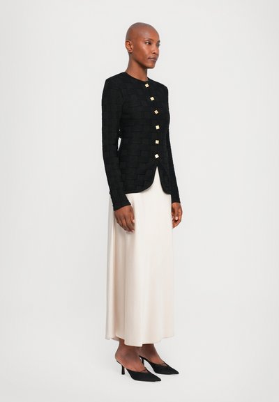 Black textured jacket with gold square buttons, paired with a cream skirt and black pointed heels. Smooth fabric and fitted design.