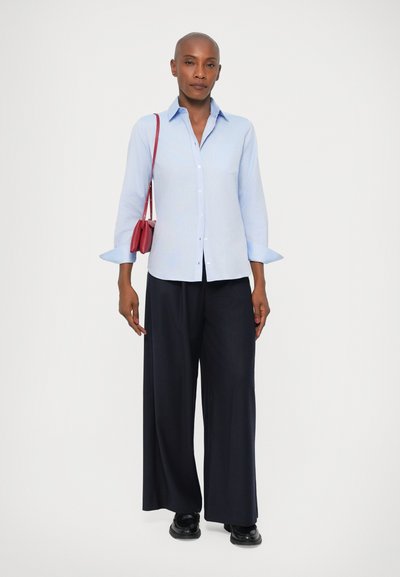 Lysblå knap-ops t-shirt med lange ærmer, en brystlomme, parret med marineblå bredbenede bukser og en lille rød crossbody taske. Sorte sko.