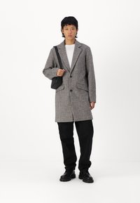 Abrigo largo de patrón de pata de gallo en gris y negro, con un cuello con muesca, dos bolsillos frontales y cierre de botón, combinado con pantalones y zapatos negros.