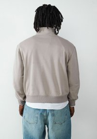 Grau Sweatshirt mit hohem Kragen, gerippten Manschetten und Saum, kombiniert mit hellblauen Jeans, von hinten vor einem neutralen Hintergrund gezeigt.