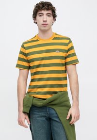 Ριγέ βαμβακερό t-shirt σε κίτρινο και πράσινο με κοντά μανίκια, στρογγυλή λαιμόκοψη και μικρό λογότυπο στο στήθος. Φορεμένο με μπλε τζιν παντελόνι.