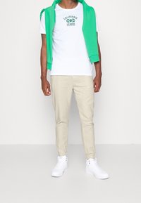 Vit t-shirt med grön "California League"-tryck, beige tapered byxor och vita hög-top sneakers. Modellen har en grön hoodie hängande över axlarna.