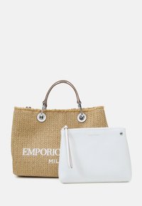 Halvstråväska med bruna handtag och präglad logotyp. Vit dragkedjesäck med silverfärgade detaljer. Båda väskorna har släta ytor.