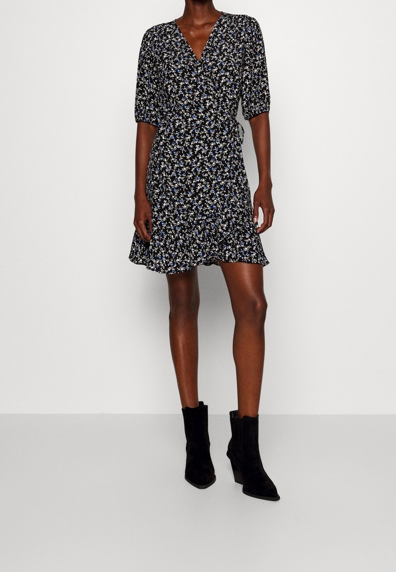 Robe portefeuille noire à motif floral avec manches courtes bouffantes, ourlet à volants, ornée de petites fleurs bleues, blanches et jaunes. Assortie à des bottines noires à la cheville.