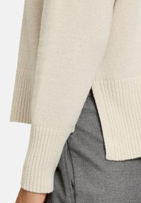 Pull en tricot beige avec des poignets et un ourlet côtelés, présentant une fente sur le côté et une texture douce, associé à un tissu gris en dessous.