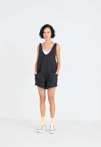 Combinaison sans manches noire avec de grandes poches avant, associée à un débardeur blanc en dessous, des chaussettes jaunes et des baskets multicolores.