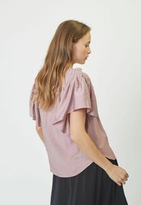 Blusa rosa chiaro con maniche fluttuanti, scollatura arricciata, tessuto testurizzato e orlo curvo, abbinata a una gonna nera.