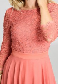Robe en dentelle corail avec un corsage en dentelle ajusté, des manches trois-quarts et une jupe fluide plissée. Ceinture lisse en accent.