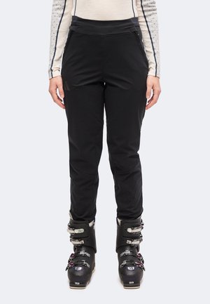 Personne portant un pantalon de ski noir, un haut crème à manches longues à manches à pois, et des chaussures de ski noires, debout face à l'avant.