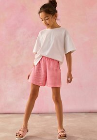 T-shirt blanc oversized associé à un short en vichy rose avec une taille élastique, porté avec des sandales rose clair sur un fond neutre.