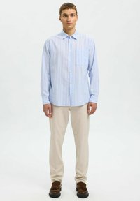 Camisa de botones a rayas azul claro con un bolsillo en el pecho, combinada con pantalones beige y zapatos marrones. La camisa tiene mangas largas y un cuello.