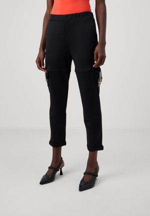 Person iført sorte taprede cargobukser med opkørte manchetter, sorte spidse hæle og en orange top, stående mod en ensfarvet grå baggrund.