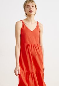 Robe rouge à niveaux sans manches avec un col en V. Fabriquée en tissu léger, avec des bretelles froncées et une texture douce.