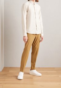 Lichtbeige langwerpige blouse met knoppen, gecombineerd met mosterdkleurige op maat gemaakte broek en witte sneakers. Eenvoudig ontwerp, gladde stofstructuur.