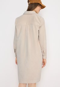 Beige kordflöjelsklänning med långa ärmar, skjortkrage och knäppte manschetter. Har en bakre axel och lätt plissering.